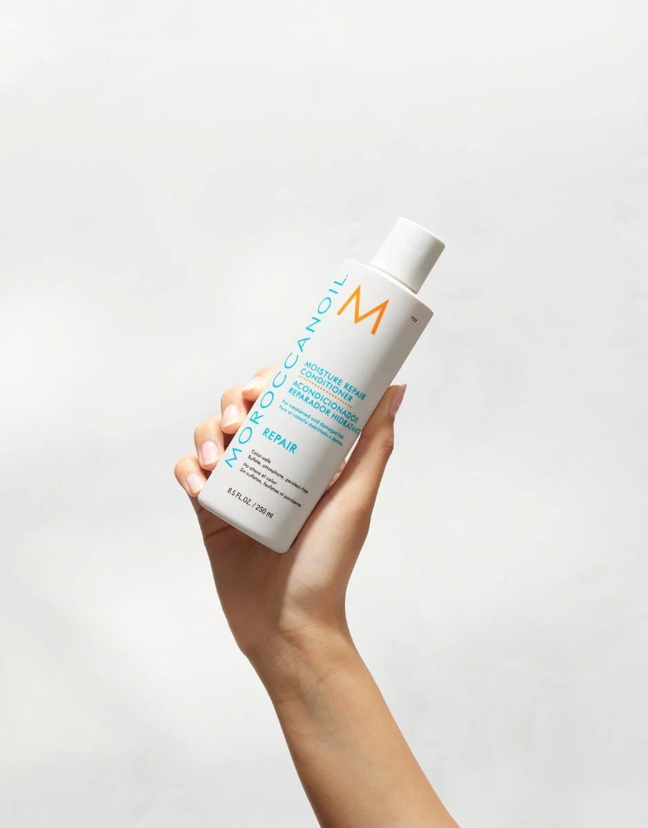 A hand holds the white 250 mL Moroccanoil Moisture Repair Conditioner bottle, showing its turquoise product label and repair line.