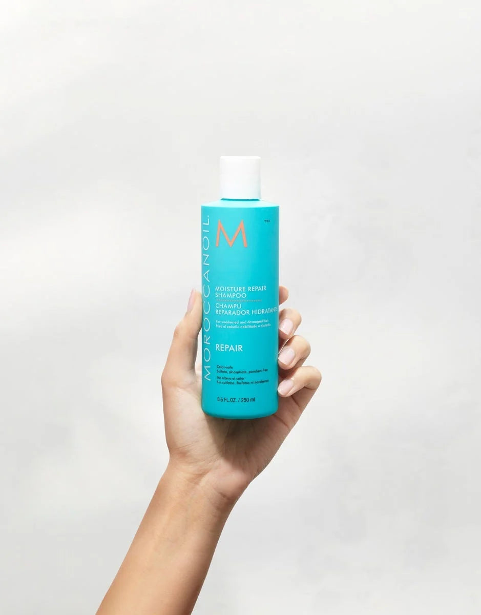 A hand holds the turquoise 250 mL Moroccanoil Moisture Repair Shampoo bottle, showing its front label and repair line.